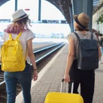 Two traveling women with suitcase backpacks walking along platform